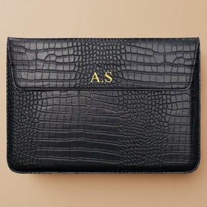 May include: Black faux leather laptop sleeve with a crocodile skin texture. The sleeve has a flap closure and gold-coloured initials "A.S" on the front. The background is a solid tan colour.