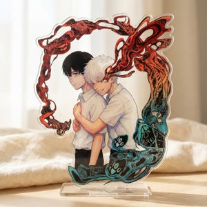 Puede incluir: Soporte acrílico con dos personajes de estilo anime abrazándose. La obra de arte está enmarcada por un borde decorativo rojo y azul. Los personajes visten camisas blancas y pantalones negros. El soporte es transparente y tiene una base rectangular.