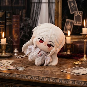 Peut inclure: Une poupée en peluche avec une fourrure blanche et des yeux rouges, vêtue d'une tenue blanche avec des détails complexes. La poupée est posée sur une table en bois avec des cartes à jouer et des bougies, créant un thème gothique ou fantastique.