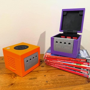 May include: An orange and gray video game console sits next to a purple console with an open lid. The purple console is stacked on top of several video game cases. The consoles are on a wooden surface.