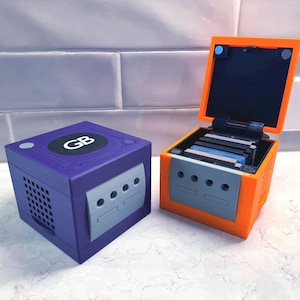 May include: Two colorful game consoles, one purple and one orange, sit side-by-side. The purple console has a gray panel with three circular openings and the letters "GB" on top. The orange console has its top open, revealing game cartridges.