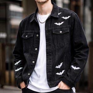 Può includere: Giacca di jeans nera con motivi di pipistrelli bianchi. La giacca ha un design classico con bottoni, tasche sul petto e colletto. I pipistrelli bianchi sono posizionati sulla parte anteriore e sulle maniche. La giacca è abbinata a jeans neri e una maglietta bianca.