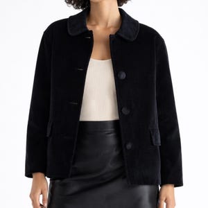May include: A black corduroy jacket with a rounded collar and three buttons. The jacket has two front pockets and long sleeves. The model is wearing a white vest top and a black skirt.