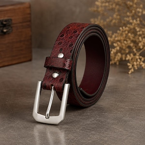 Op de afbeelding: Een donkerrode riem met een textuur met stippenpatroon die doet denken aan struisvogelleder. De riem is voorzien van een gepolijste zilverkleurige metalen gesp en twee zilverkleurige metalen klinknagels bij de lus, opgerold in een cirkelvorm.