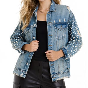 Puede incluir: Chaqueta vaquera azul claro con detalles desgastados y adornos de perlas en las mangas. La chaqueta tiene un cuello clásico, cierres de botón y bolsillos delanteros. Las mangas están adornadas con numerosas perlas blancas, añadiendo un toque de elegancia al denim casual.