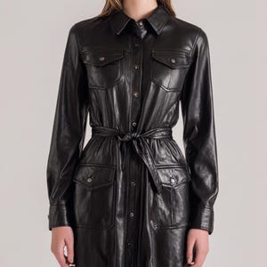 Puede incluir: Un vestido de cuero negro con mangas largas, cuello camisero y cierre de botones. El vestido presenta dos bolsillos en el pecho, dos bolsillos en las caderas y un cinturón a juego atado a la cintura. El vestido es de cuero liso y brillante.