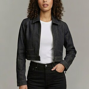 Puede incluir: Chaqueta vaquera azul oscuro con cremallera, cuello y bolsillos. La chaqueta está recortada en la cintura y se usa con una camiseta blanca y unos vaqueros negros. La modelo tiene el pelo rizado.