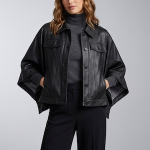 Puede incluir: Chaqueta de cuero negra con un diseño único tipo capa. La chaqueta presenta un cuello, dos bolsillos en el pecho y cierres de botón. La modelo lleva un cuello alto gris y pantalones negros.