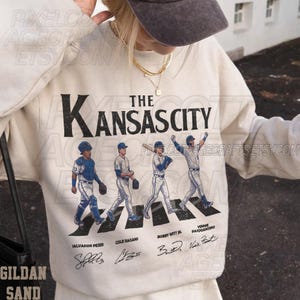May include: Cream-colored sweatshirt featuring the text "The Kansas City" above an illustration of four baseball players crossing a crosswalk. The names Salvador Perez, Cole Ragans, Bobby Witt Jr., and Vinnie Pasquantino are below the players.