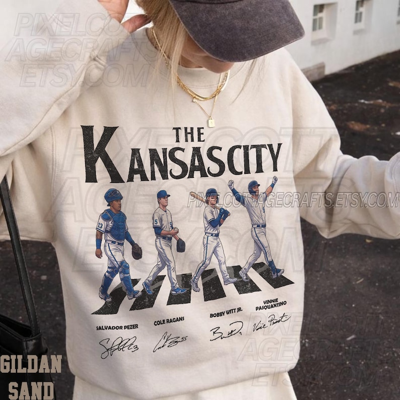 May include: Cream-colored sweatshirt featuring the text "The Kansas City" above an illustration of four baseball players crossing a crosswalk. The names Salvador Perez, Cole Ragans, Bobby Witt Jr., and Vinnie Pasquantino are below the players.