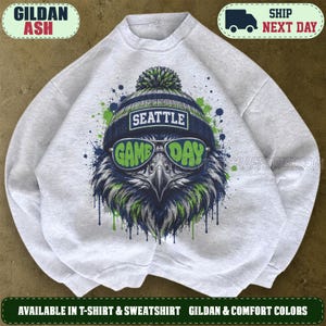 May include: A light gray crewneck sweatshirt featuring a graphic of an eagle wearing a blue and green knit beanie and sunglasses. The text SEATTLE is printed on the beanie, and the text GAME DAY is printed across the lenses of the sunglasses.