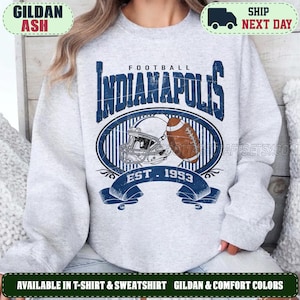 Puede incluir: Una sudadera gris claro de cuello redondo con un estampado gráfico azul oscuro que dice Indianapolis Football Est. 1953. El diseño incluye un casco de fútbol americano y un balón de fútbol marrón centrados en un óvalo con una cinta azul.