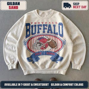 May include: A sand-colored crewneck sweatshirt featuring a distressed graphic print. The design includes the text "FOOTBALL BUFFALO" in blue, a football helmet, a football, and a blue banner with the text "EST. 1960" centered on the chest.