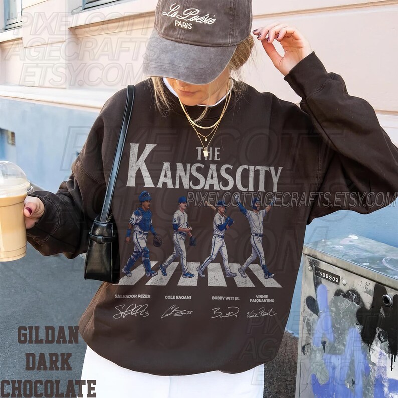 May include: Dark chocolate-colored sweatshirt featuring a graphic of Kansas City baseball players crossing a street, with player names below. The sweatshirt has the text "The Kansas City" in white. The brand name "Gildan" is visible at the bottom.