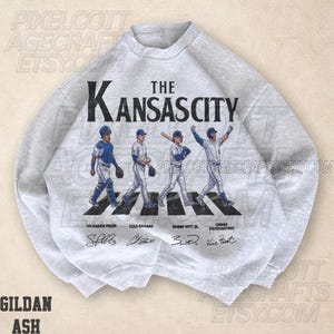 May include: A light gray sweatshirt featuring the text "The Kansas City" above an illustration of four baseball players crossing a crosswalk. The names and signatures of the players are below the illustration.