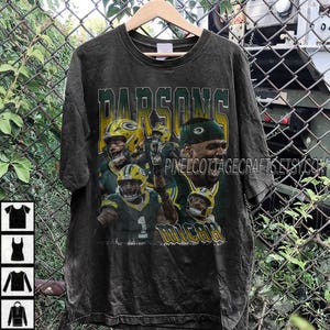 May include: A black t-shirt featuring a distressed design with the name "Parsons" in green and yellow lettering. The shirt showcases images of football players in green and yellow uniforms. The word "Micah" is also visible.