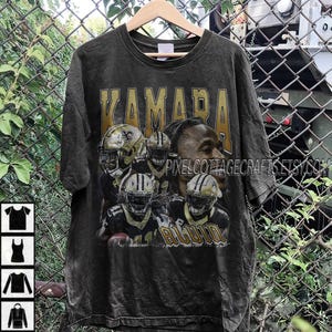 Peut inclure: T-shirt noir avec un graphisme vieilli d'un joueur de football américain et le mot "KAMARA" en lettres dorées. Le t-shirt comprend également le mot "ALVIN" en lettres dorées. Le t-shirt est suspendu à un cintre en bois.