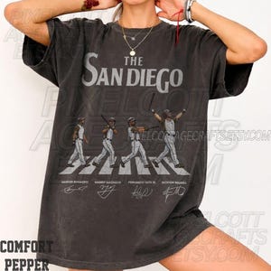 Può includere: T-shirt grigio scuro con la scritta "The San Diego" e quattro giocatori di baseball che attraversano un passaggio pedonale, con nomi e firme dei giocatori. La maglietta ha un aspetto vintage.