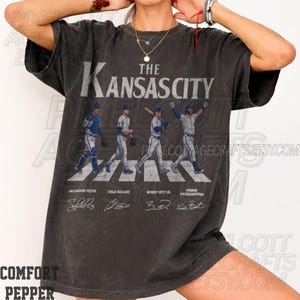 May include: Dark gray t-shirt featuring the text "The Kansas City" above an image of four baseball players crossing a crosswalk. The players' names and signatures are below the image.
