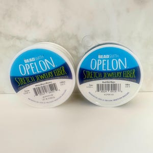 May include: Two spools of Beadsmith Opelon stretch jewelry fiber. One spool is white, the other is black. Each spool has a diameter of 7mm (0.028 in) and contains 100m (328 ft) of fiber. The packaging is white with blue and black text.