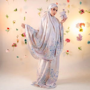 May include: A light-colored prayer garment set with a floral pattern, including a head covering and matching dress. The fabric drapes gracefully, and the design features soft blues and purples. The person is holding a small, decorated box.