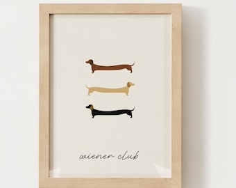 May include: Framed art print featuring three dachshund illustrations in brown, tan, and black. The phrase "wiener club" is written in a cursive font below the dogs. The artwork is set against a cream background and framed in a light wood.