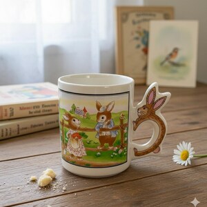 Peut inclure: Tasse en céramique blanche avec une illustration de lapin et une anse en forme de lapin. La tasse présente une scène champêtre avec deux lapins près d'une clôture. Une marguerite et des livres sont en arrière-plan.