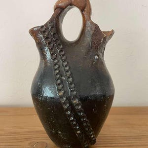 以下が含まれることがあります： ユニークなハンドルと装飾的なビーズのアクセントが付いた、手作りの濃い茶色のセラミック花瓶。 花瓶は球根状のボディとテクスチャード加工された表面を持ち、素朴な美学を示しています。 ハンドルには円形の開口部があります。