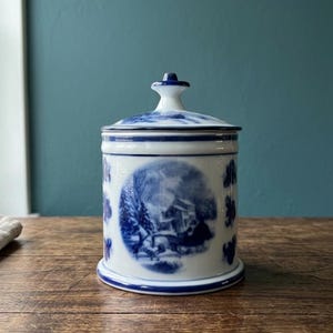 Puede incluir: Un tarro de cerámica blanco y azul con tapa. El tarro presenta una escena de paisaje azul y blanco y detalles florales. El tarro es cilíndrico con una base redondeada y un pequeño pomo en la tapa.