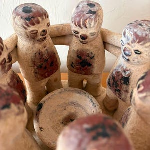 May include: A circular clay sculpture featuring a group of stylized figures standing in a ring with arms linked around each other. The figures have a textured, light tan surface with dark brown and black painted accents on their heads and torsos.