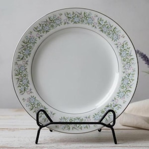 Puede incluir: Un plato de porcelana blanco con un borde floral en tonos azules, verdes y rosas, acentuado con un borde plateado. El plato se exhibe en un soporte de metal negro, mostrando su elegante diseño. Ideal para servir comidas o como pieza decorativa.