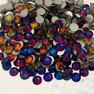 May include: A large assortment of iridescent glass rhinestones in various shades of blue, purple, orange, and yellow. The rhinestones are scattered on a white surface.