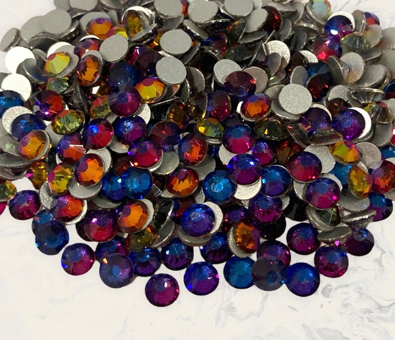 May include: A large assortment of iridescent glass rhinestones in various shades of blue, purple, orange, and yellow. The rhinestones are scattered on a white surface.