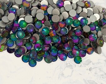 May include: A large pile of iridescent green, purple, and blue rhinestones. The rhinestones are scattered on a white surface.