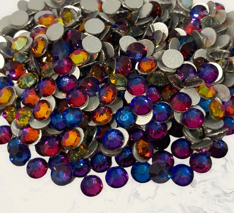 May include: A large pile of iridescent glass rhinestones in shades of blue, purple, orange, and red. The rhinestones are flat-backed and have a silver backing.