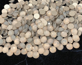 May include: A collection of iridescent, round-cut, opal-colored gemstones. The gemstones have a faceted, domed top and a flat, silver-colored backing. The gems are scattered, creating a textured, sparkling effect.