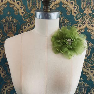 May include: A light green fabric flower with multiple layers and a cluster of small white beads in the center. The flower is pinned to a mannequin. The background is a teal wallpaper with a gold and purple paisley design.
