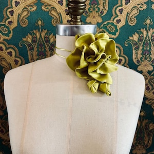 Puede incluir: Un broche de flores de satén verde chartreuse se exhibe en un maniquí. La flor tiene múltiples capas con volantes y está unida a una fina cinta dorada. El maniquí se encuentra sobre un fondo estampado en verde azulado y dorado.