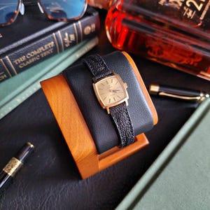 Pode incluir: Um relógio Omega dourado com mostrador retangular e pulseira de couro preto texturizado. O relógio é exibido em um suporte de madeira e couro preto. O mostrador do relógio tem ponteiros e marcadores dourados. Um relógio clássico.