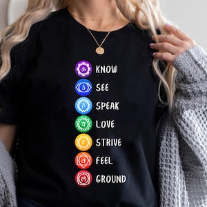 May include: Black t-shirt featuring a colorful chakra design with the words "KNOW, SEE, SPEAK, LOVE, STRIVE, FEEL, GROUND" in white. The design includes symbols and colors associated with each chakra.