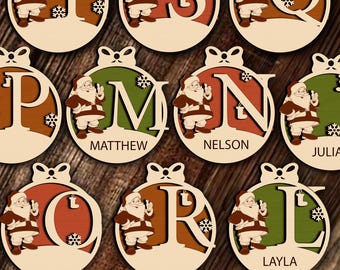 Pode incluir: Enfeites de Natal de madeira em forma de bola, cada um com uma letra e uma figura do Pai Natal. As letras são de várias cores, com nomes como Matthew, Nelson e Layla. O texto "CHRISTMAS BALLS" e "LASER CUT FILE" está na parte inferior.