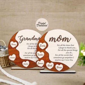 Puede incluir: Dos placas de madera, una con la palabra "Abuela" y la otra "Mamá". Cada placa tiene un recorte en forma de corazón con nombres y un mensaje sincero. El diseño presenta un color de madera claro con un acento de madera más oscuro.