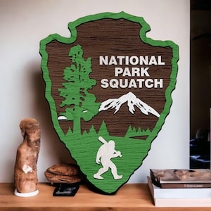 May include: Wooden sign in the shape of an arrowhead with the words "NATIONAL PARK SQUATCH". The sign features a green border, a tree, a mountain, and a silhouette of Bigfoot. The sign is brown and green.