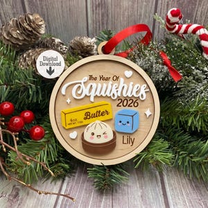 Può includere: Un ornamento rotondo in legno con il testo The Year Of Squishies 2026 e il nome Lily. Include icone in stile 3D di un panetto di burro giallo da 113g, un cubetto di ghiaccio blu con una faccia e un raviolo in una ciotola che fa l'occhiolino.