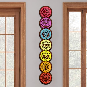 May include: A vertical arrangement of seven circular wooden chakra wall art pieces. Each piece features a different color and symbol, including pink, purple, blue, yellow, orange, and red. The wooden frames have a dark brown finish.