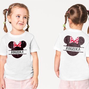 May include: White t-shirt featuring a Minnie Mouse design with a red polka dot bow and the name "Angelica" in black. The design is on the front and back of the shirt. The child is wearing pink pants.