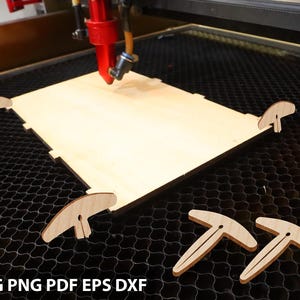 Op de afbeelding: Een lasersnijder die werkt aan een lichtgekleurd houten project. De rode nozzle van de machine bevindt zich boven het hout, met verschillende kleine, T-vormige houten stukken in de buurt. De tekst "SVG PNG PDF EPS DXF" is zichtbaar.
