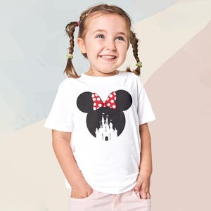 May include: A white t-shirt featuring a black silhouette of Minnie Mouse's head with a red and white polka dot bow. The silhouette includes a castle design. The shirt is worn by a young girl.