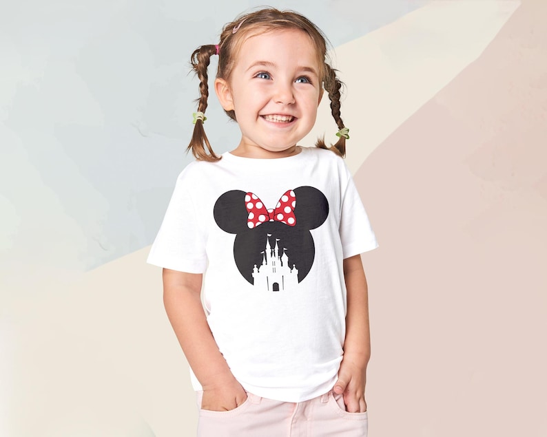 May include: A white t-shirt featuring a black silhouette of Minnie Mouse's head with a red and white polka dot bow. The silhouette includes a castle design. The shirt is worn by a young girl.