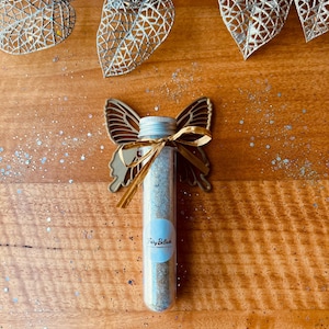 May include: A clear glass tube filled with a light-coloured substance, decorated with a gold butterfly-shaped embellishment and a gold ribbon. The tube has a silver cap and a label that reads "Fairy Belish". The background features decorative gold leaves and glitter.